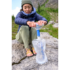 Vestida con equipo para actividades al aire libre, una persona utiliza el sistema de filtración de agua Botella Flexible Platypus Platy 2L en un terreno rocoso, rodeada de árboles verdes y terreno accidentado, capturando un espíritu aventurero.