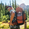 Una persona con una camiseta estampada y gafas de sol se encuentra en una zona montañosa boscosa, llevando una mochila naranja en la que se ve una botella de agua flexible Platypus Platy de 2 l. Los pinos y la cima de una montaña crean un idílico telón de fondo al aire libre.