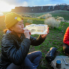 Una persona con un gorro amarillo y una chaqueta negra bebe de una botella Platypus SoftBottle de 1 l mientras está sentada al aire libre al atardecer, con colinas cubiertas de hierba detrás. Cerca de allí, otra persona con una chaqueta roja está sentada entre equipos de acampada dispersos.