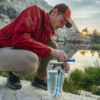 De rodillas junto a un lago de montaña, con una chaqueta roja y una gorra naranja, una persona utiliza un filtro de agua portátil para purificar el agua de su Bolsa de Hidratación Platypus Big Zip EVO. El paisaje rocoso se refleja en el cielo despejado.