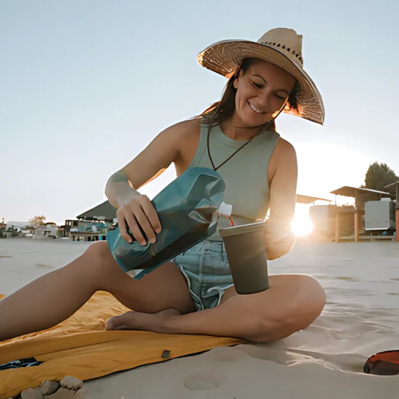 Una mujer con sombrero para el sol y camiseta sin mangas sonríe mientras sirve un trago de su botella de vino Platypus PlatyPreserve de 0,8 l en una taza mientras se relaja sobre una tela amarilla en la playa. El sol poniente proyecta un cálido resplandor con edificios y árboles al fondo.