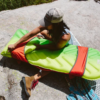 Una persona está sentada sobre una roca, ajustando una colchoneta verde y roja para dormir junto a una férula para postes de carpa MSR de Tubo de Reparación. Lleva una gorra, gafas de sol y botas. Debajo de ella hay una tela a cuadros, con césped y plantas de fondo.