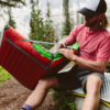 Una persona con una camisa y una gorra rojas se sienta en un banco al aire libre, cerrando la cremallera de un saco de dormir rojo y verde con una férula para postes de carpa Tubo de Reparación MSR Tent Pole Repair Splint (copia) (copia) a su lado. Árboles altos y un paisaje borroso crean un fondo tranquilo.