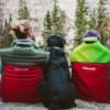 Dos personas con ropa de exterior se sientan en sillas ThermaRest con un perro negro entre ellas, frente a unos pinos altos en un entorno tranquilo. Cerca de allí, una férula de reparación de postes de carpa MSR de Tubo de Reparación asoma desde una mochila, lista para cualquier desafío de aventura.