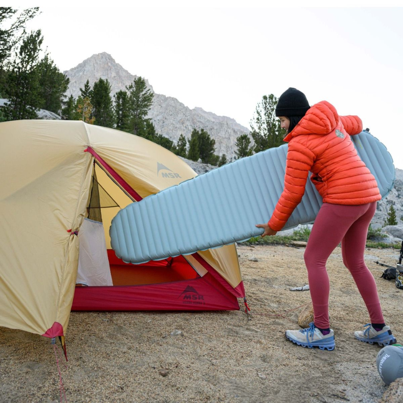 En una zona montañosa, alguien con chaqueta roja y pantalón rosa monta una tienda de campaña. Lleva una colchoneta Therm-a-Rest NeoAir XLite NXT, de color azul claro, hacia la entrada de la tienda. El suelo arenoso está enmarcado por árboles y colinas rocosas de fondo.