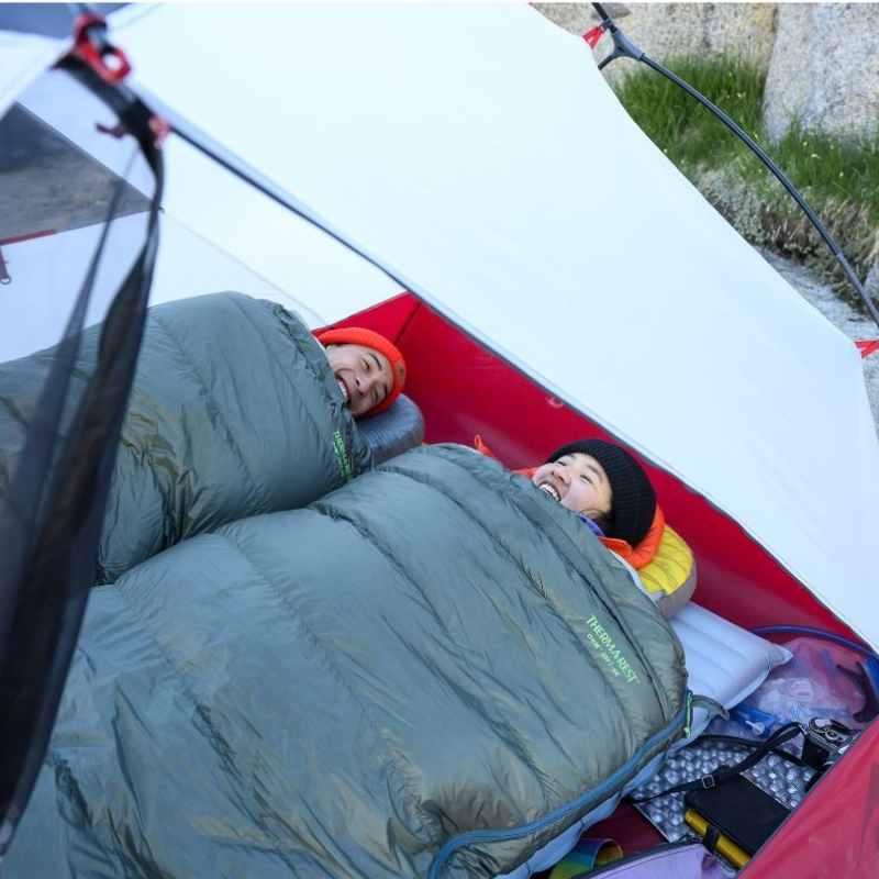 Dos personas en sacos de dormir, amortiguados por su Colchoneta Therm-a-Rest NeoAir XTherm NXT (copia), sonríen mientras están acostados uno al lado del otro en una acogedora tienda de campaña con equipo y artículos personales dispersos; la tienda está instalada al aire libre en una zona rocosa.