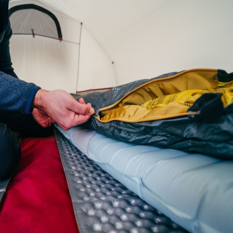 Una persona cierra la cremallera de un saco de dormir dentro de una tienda de campaña, que presenta una acogedora colchoneta Therm-a-Rest NeoAir XTherm NXT gris con una capa roja debajo. El acogedor interior crea un ambiente ideal para acampar.