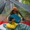 Dentro de una tienda de campaña, una persona de pelo rizado con un gorro marrón infla una almohada de camping azul mientras su Kit de Reparación Therm-a-Rest Instant Field (copia) reposa cerca. Están sentados en un saco de dormir amarillo, rodeados de un frondoso bosque. La tienda de campaña parcialmente abierta revela el sereno paisaje exterior.