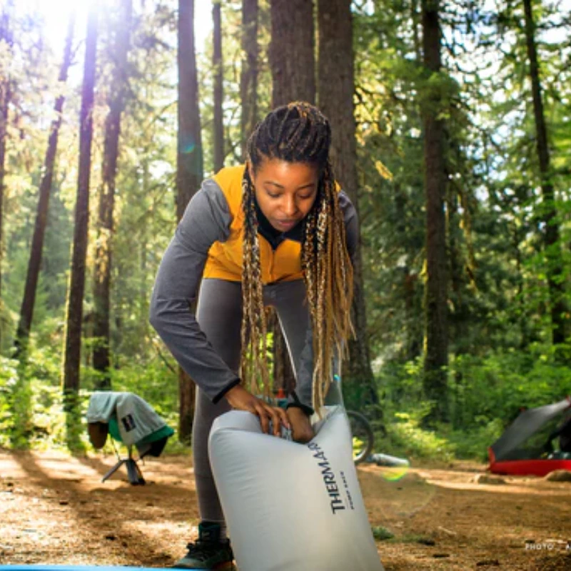 Una persona con el pelo largo trenzado, con un chaleco amarillo y calzas grises, prepara su Colchoneta Therm-a-Rest NeoAir XTherm NXT MAX (copia) en el suelo del bosque. La luz del sol se cuela entre los árboles, con una tienda de campaña y una bicicleta al fondo.
