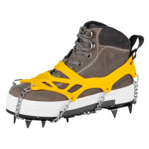Una bota de senderismo marrón con cordones grises está equipada con un dispositivo de tracción amarillo que cuenta con clavos de metal y cadenas para un mejor agarre, similar a los Guantes Grivel Rock Gloves. El dispositivo se ajusta perfectamente a la bota, ideal para terrenos helados o nevados.