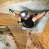 Un escalador con camiseta negra y casco Grivel Salamander 2.0 (copia) asciende por una pared rocosa con textura, agarrándose con la mano derecha a un punto de apoyo mientras con la izquierda se agarra a una grieta. El equipo de escalada está sujeto a su arnés.