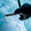 Una mano enguantada utiliza con destreza un tornillo para hielo Petzl Laser Speed (copia) para perforar el hielo azul y capturar las intrincadas texturas de los fragmentos de hielo y el metal. Las formaciones de hielo de textura suave en el fondo crean un contraste visual sorprendente.