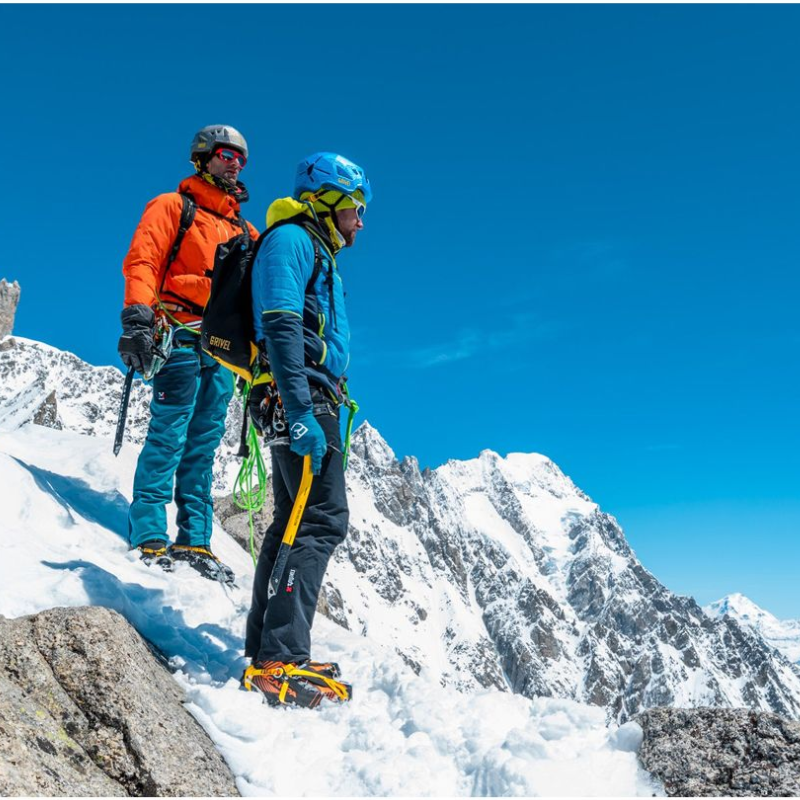 Dos escaladores con ropa de invierno se encuentran en la cima de una montaña nevada, sosteniendo un Piolet Clásico Grivel G1 (copia) y mirando a lo lejos. Ambos llevan cascos y equipo de escalada, mientras los picos cubiertos de nieve se recortan contra un cielo azul claro.
