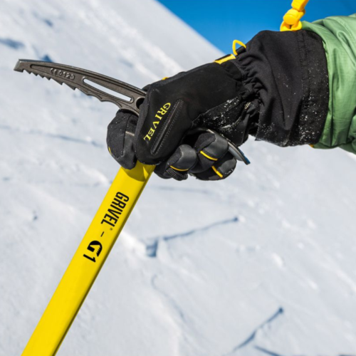 Una persona sostiene un piolet amarillo Piolet Clásico Grivel Nepal S.A. (copia) con una mano enguantada, clavándolo en la nieve. Sus guantes tienen detalles en negro y amarillo que complementan su chaqueta verde contra el fondo del cielo azul claro.