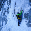 Un escalador con chaqueta verde, casco y crampones asciende por una pared rocosa cubierta de hielo. Equipado con piolets The Light Machine de Grivel y una cuerda para mayor seguridad, avanza por la nieve y el hielo del terreno montañoso, duro y frío.