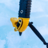 Primer plano de un mango de piolet amarillo y negro, similar al de las Barras para Crampones Grivel Valter Long Bar (copia), sobre un suelo nevado. Presenta una textura de agarre con una correa negra adjunta para asegurar la herramienta.