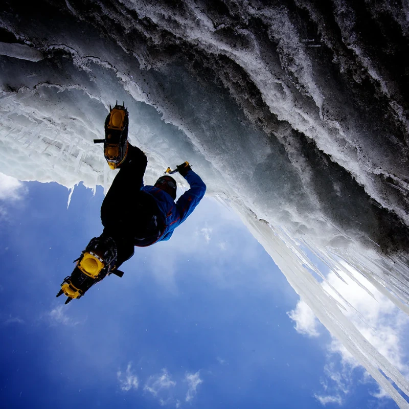 Con verdadera destreza de montañismo técnico, un escalador agarra los crampones para montañismo técnico Grivel G14 contra la pared helada. Su silueta se ve acentuada por el cielo y las nubes, resaltando la pendiente y la euforia de este desafiante ascenso.