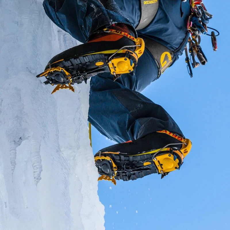 Un escalador con botas amarillas y negras y crampones de escalada mixtos Grivel G20 Plus (copia) asciende por un terreno helado. Los piolets y el equipo cuelgan de su cintura contra un cielo azul despejado como telón de fondo.
