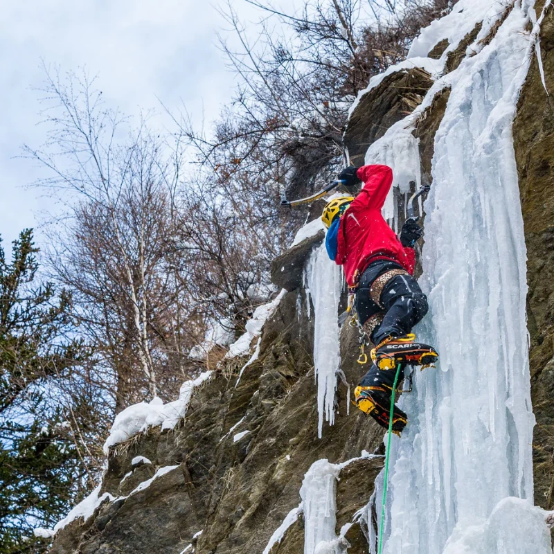 Con una chaqueta roja, un escalador escala con piolets una cascada helada en un acantilado rocoso, asegurado con una cuerda verde y crampones de escalada mixta Grivel G20 Plus (copia). La nieve y las ramas desnudas se destacan contra el cielo nublado.