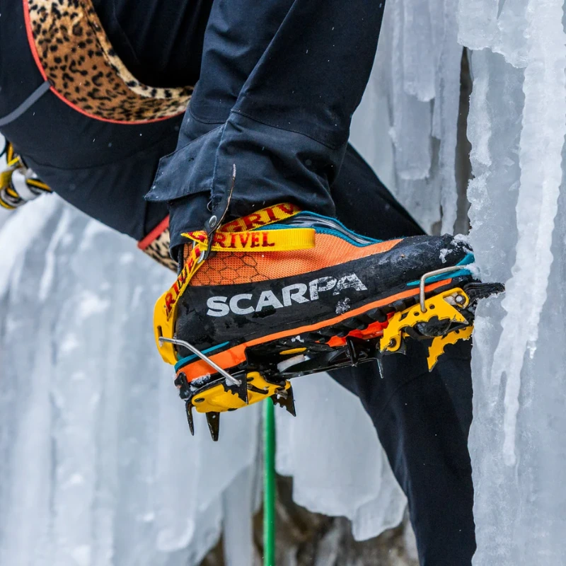Primer plano de una bota de escalador con crampones de escalada mixtos Grivel G20 Plus (copia) agarrando superficies heladas. La bota vibrante muestra su marca mientras se coloca un arnés de escalada en medio de formaciones de hielo en cascada.