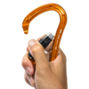 Una mano sostiene un mosquetón Grivel S1G Steel One Twin Gate naranja con dispositivo de seguridad integrado para escalada, con forma de gancho y mecanismo de bloqueo negro. El pulgar presiona el bloqueo, lo que resalta su diseño de doble gatillo para mayor seguridad.