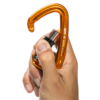 Una mano sostiene un mosquetón de seguridad Grivel de color naranja con mecanismo de cierre negro. El mosquetón está abierto, mostrando su gatillo con resorte, y la marca "Grivel" es visible en el costado.