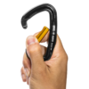 Una mano sostiene un elegante mosquetón Grivel K1 negro con gatillo naranja. Cuenta con un mecanismo de cierre seguro y una marca sutil en el lateral, mientras que los dedos lo agarran con cuidado para abrirlo o asegurarlo.
