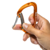 Una mano sostiene un mosquetón de escalada Grivel K8G Sigma naranja con gatillo plateado, colocado entre el pulgar y el índice para resaltar su forma ergonómica y su construcción metálica.