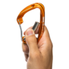 Una mano sostiene un mosquetón de escalada Grivel K8G Sigma naranja con gatillo de alambre, ligeramente abierto para revelar la marca "GRIVEL" en su costado sobre un fondo blanco liso.