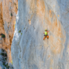 Vestido con una camisa verde y pantalones naranjas, un escalador escala una pared rocosa empinada y grande asegurada con equipo de escalada Cinta Express Grivel All-Round Alpha y cuerdas. Usa un casco para mayor seguridad. La roca gris tiene manchas naranjas, con vegetación escasa que agrega color al fondo.