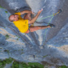 Con una camiseta amarilla y pantalones cortos de color naranja, un escalador trepa un acantilado rocoso empinado con las manos pintadas de tiza. Confiando en su equipo Grivel, está firmemente sujeto a la pared con la Pack Cinta Express Grivel All-Round Alpha. Una caída vertical y una vegetación distante se extienden hacia el fondo.