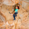 Una mujer con una camiseta verde y unos vaqueros azules está escalando una roca en un acantilado de color marrón anaranjado, equipada con un Pack Cinta Express Grivel Sport Stealth. Sostiene la roca con la mano derecha, sujeta con una cuerda de seguridad. Con el pelo atado hacia atrás, parece concentrada y decidida.
