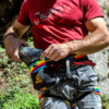 En un entorno rocoso al aire libre, alguien con una camisa roja y pantalones de escalada grises ajusta un arnés de escalada colorido mientras sostiene la Bolsa Magnesera Grivel Trend Chalk Bag, resaltando la última tendencia.