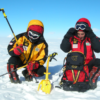 Dos personas con ropa de invierno roja y amarilla se agachan en un paisaje nevado, ajustándose las gafas. Rodeados de mochilas, sostienen un picahielos Martillo Grivel Thor (copia) con su mango amarillo brillando bajo el cielo azul claro.