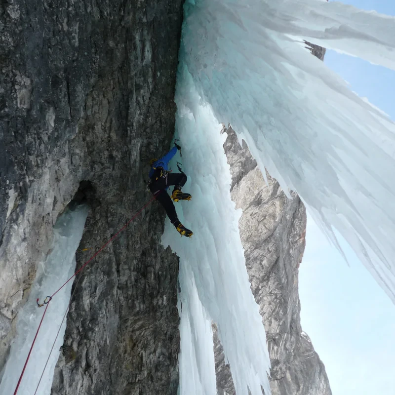 Un escalador asciende por una pared rocosa cubierta de hielo, manejando con destreza un Piolet Técnico Grivel Air Tech Evolution (copia). Sujeto con una cuerda de escalada roja, con crampones y una chaqueta azul, avanza entre grandes carámbanos que se recortan contra el cielo despejado.