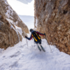Equipado con un casco y una Mochila de Montaña Grivel Spartan 45 (copia), un esquiador recorre un estrecho paso de montaña cubierto de nieve flanqueado por acantilados rocosos bajo un cielo brillante que contrasta con el accidentado terreno.