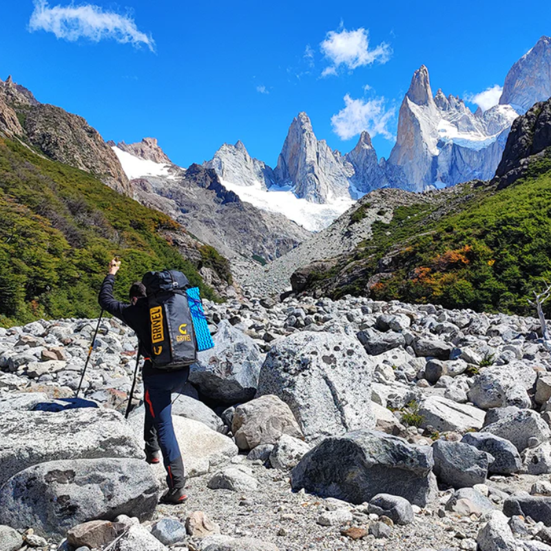 Con una Mochila de Montaña Grivel Alpine Pro 40 + 10 (copia), una persona se desplaza por terrenos rocosos, rodeados de picos escarpados y un cielo azul vibrante. Los parches de nieve adornan las montañas, mientras que la exuberante vegetación prospera debajo, resaltando la belleza contrastante de la naturaleza.