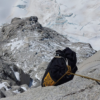 Una cuerda de escalador y una Mochila de Transporte Grivel Grand Cap 40 (copia) están aseguradas a una cornisa rocosa en una montaña cubierta de nieve, con vista a una caída pronunciada hacia un terreno accidentado y helado que se encuentra debajo.