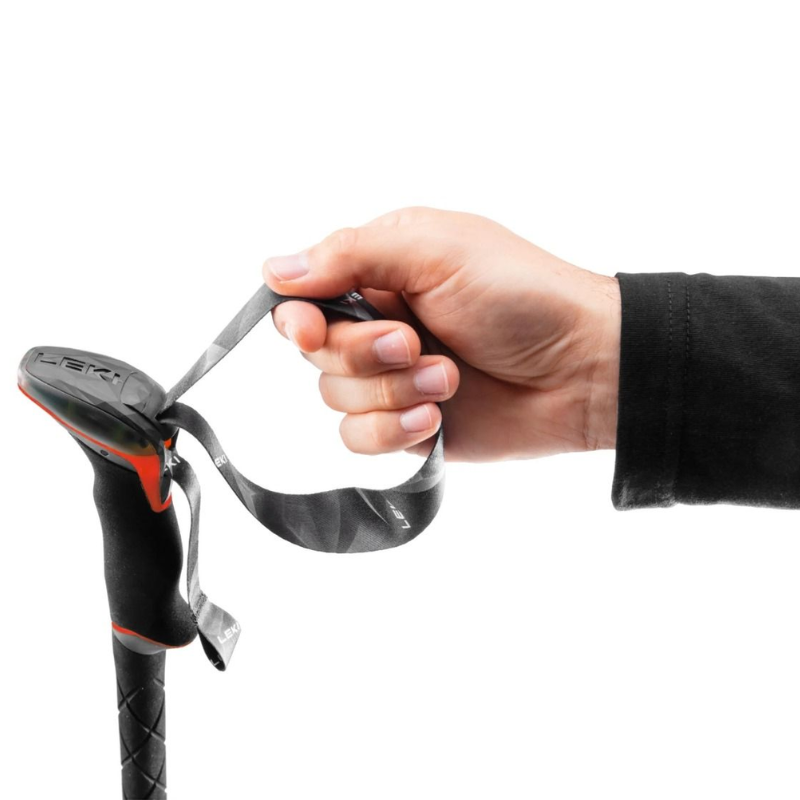 Una mano sujeta la correa de un bastón negro y rojo, similar a los Bastones de Trekking Leki Cressida Mujer (copia), sobre un fondo blanco liso. La persona lleva una camisa negra de manga larga.