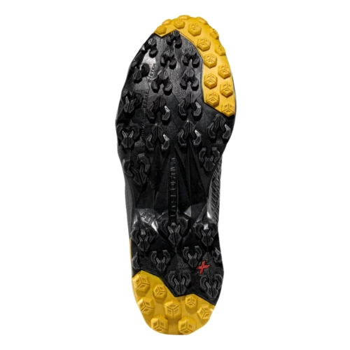 La imagen muestra la parte inferior de una zapatilla de montaña La Sportiva Akyra II GTX, que presenta un dibujo de la banda de rodadura con tacos de goma multidireccionales negros y amarillos para un agarre en diversos terrenos y un pequeño símbolo de cruz roja cerca del área superior del talón.