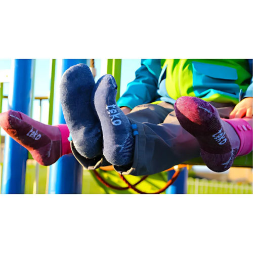 Los niños que llevan calcetines coloridos Calcetín Teko Hiking 3994 Merino XC Midweight Niños en azul, rosa y morado, combinados con botas rojas y chaquetas verdes y azules, juegan alegremente en el soleado patio de recreo.