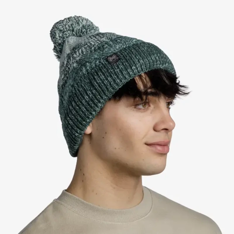 Una persona con cabello corto y oscuro lleva un gorro de lana gris y verde de la marca Gorro Buff Knitted y Polar Kim (copia), con un pompón, mientras mira hacia la derecha. Su expresión es neutra, combinada con un suéter beige sobre un fondo blanco liso.