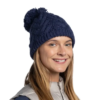 Una persona con cabello largo sonríe con un gorro azul Gorro Buff Knitted y Polar Blein (copia), con un pompón, y una chaqueta con cremallera gris claro sobre un fondo blanco liso.