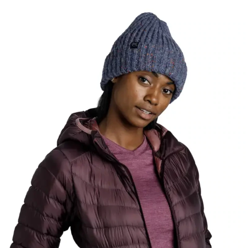 La persona, que lleva un gorro Buff Knitted y Polar Caryn (copia), una chaqueta acolchada color granate y una camisa violeta, posa con una leve sonrisa y la cabeza ladeada. La chaqueta está desabrochada para revelar la camisa que hay debajo sobre un fondo blanco.