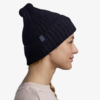La persona con trenza lleva un gorro de lana Gorro Buff CrossKnit Solid (copia) oscuro y un suéter claro. Vista de perfil, sonríe levemente, lo que le suma estilo al fondo blanco liso.