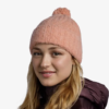 Una persona con cabello largo y rubio sonríe, vistiendo un Gorro Buff Knitted Beanie Nerla adornado con un pompón y una chaqueta oscura y acolchada sobre un fondo blanco liso.