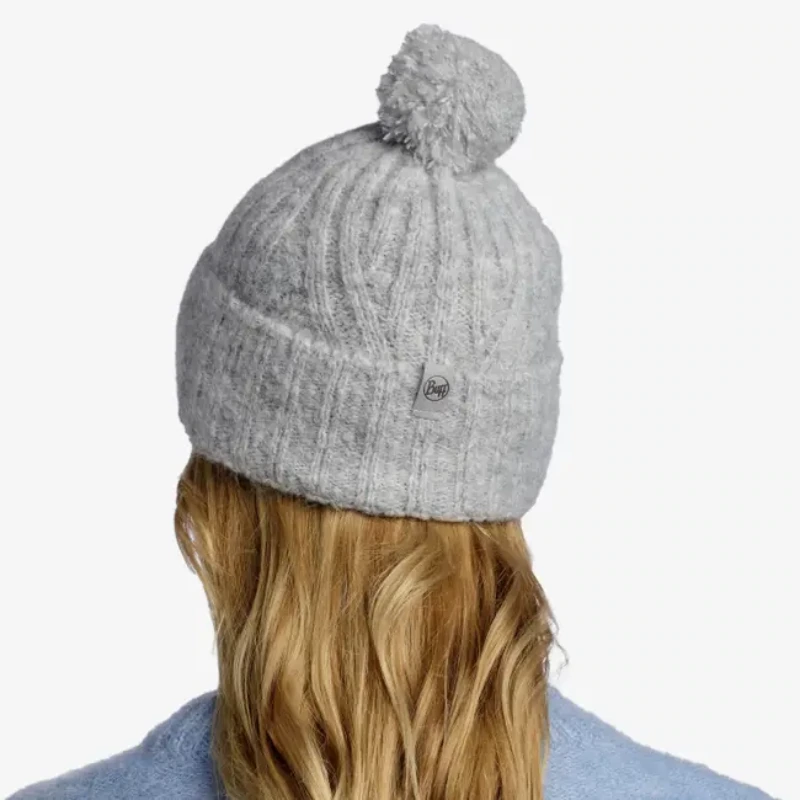 Vista desde atrás, una persona con cabello rubio largo y ondulado lleva un gorro de punto Nerla de color gris con un pompón. Su cabello cae sobre un suéter azul claro sobre un fondo blanco.