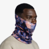La persona lleva una bandana Buff ThermoNet Solid Ultramarine y una polaina para el cuello con estampado geométrico en azul y rosa. Su cabello corto y oscuro y su camiseta beige se destacan sobre un fondo blanco liso, lo que enfatiza su estilo.