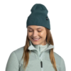 La persona, que lleva un gorro de punto Buff Knitted Beanie Lilon de color verde azulado y una chaqueta gris claro con cierre, sonríe con los ojos cerrados e inclina ligeramente la cabeza hacia abajo. El fondo es blanco liso.
