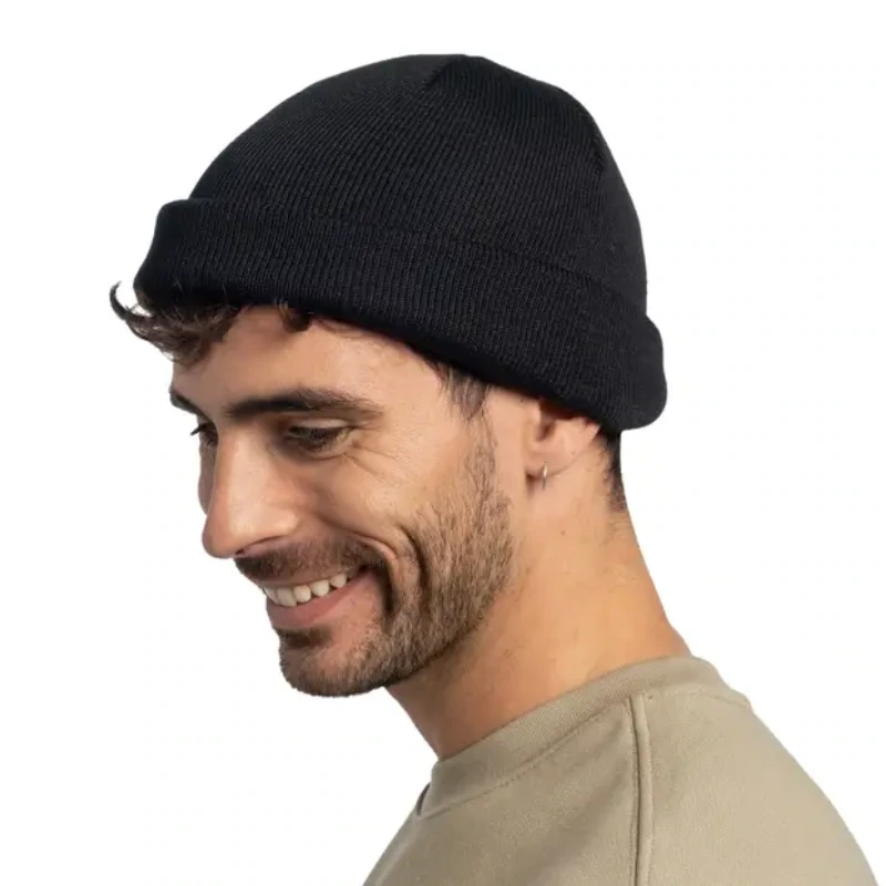 Un hombre con barba y un pendiente sonríe mientras lleva un gorro de punto y un jersey de color claro. La imagen está tomada desde un ángulo lateral sobre un fondo blanco liso.
